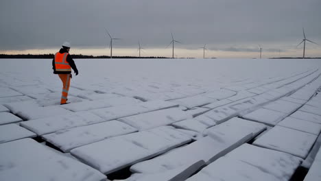 person walks on a snowy, frozen landscape with wind turbines in the distance