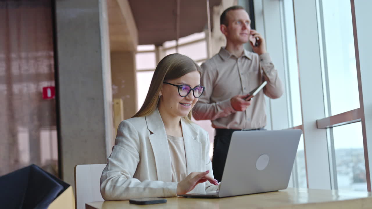 Happy smiling lady works on her laptop at the desk. Man having conversation on the mobile phone at the backdrop.