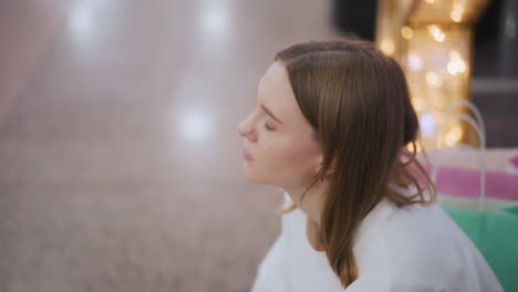 sad woman gazing around thoughtfully in a softly lit mall setting with blurred golden lights in the background, shopping bags in subtle hues complement the subdued atmosphere