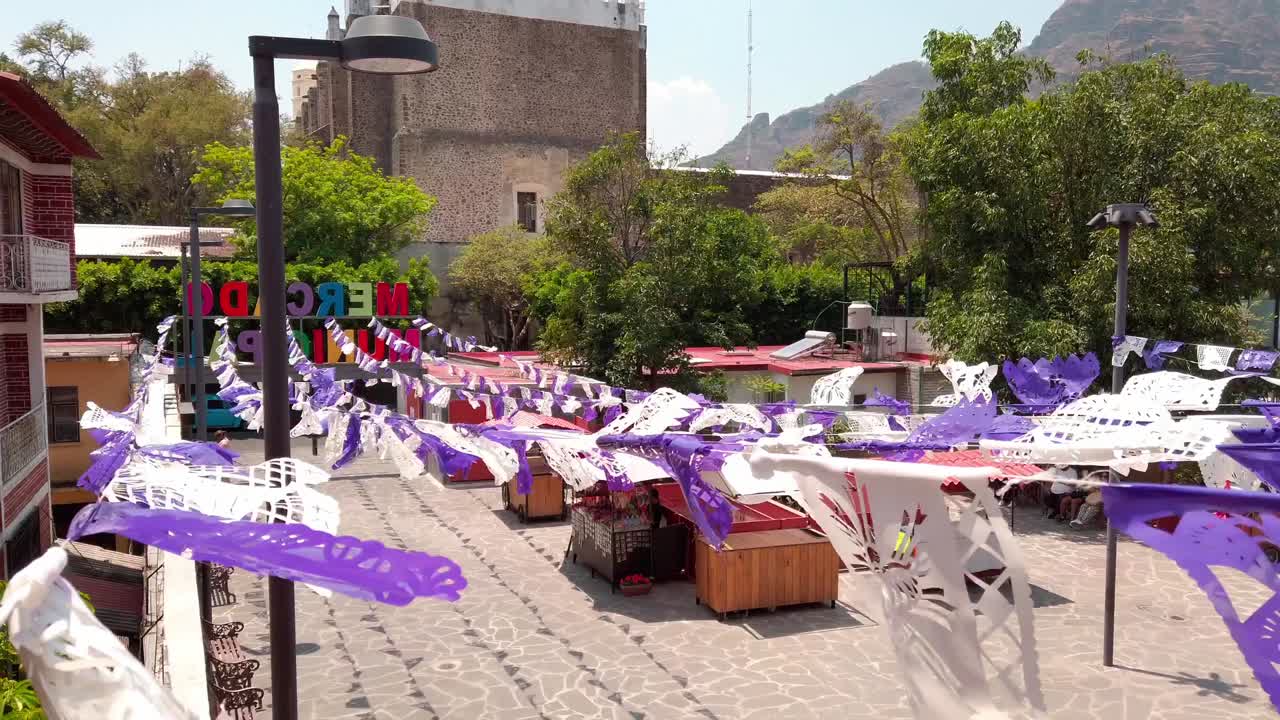 Colorful papel picado over market stalls in Tepoztlán city center with church and Tepoztlán sign in background. Static shot