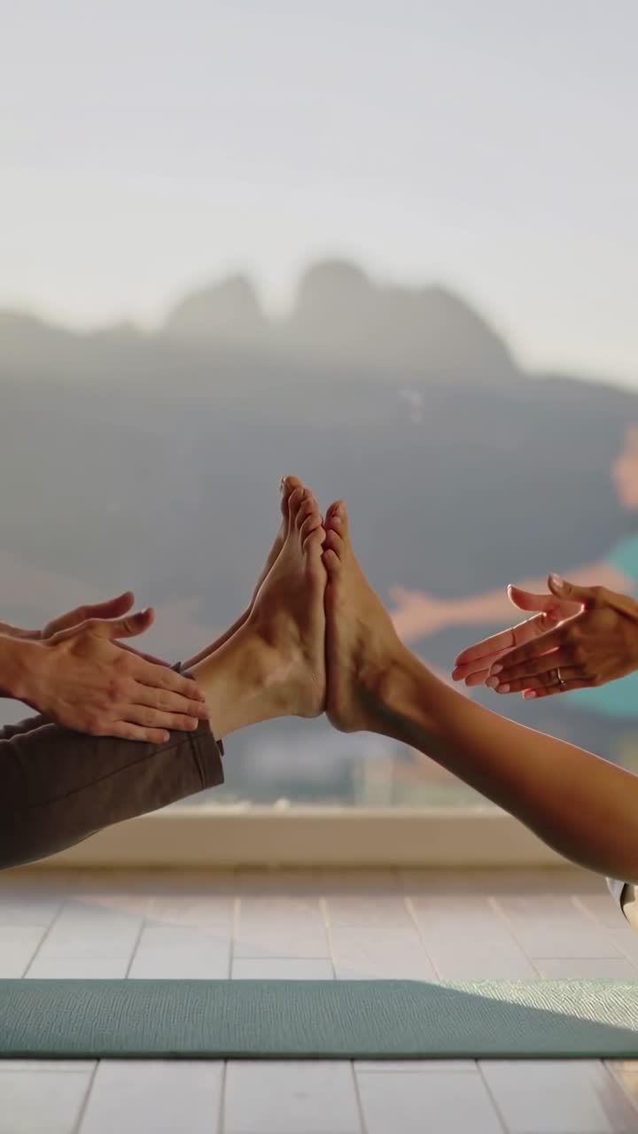 Video vertical: Iniciando estiramiento, pareja en ropa deportiva apoyando los pies en colchonetas junto a la ventana, usando las manos.