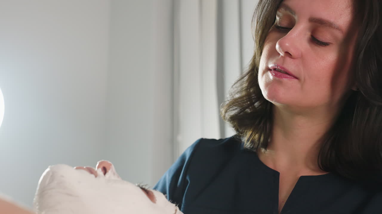 Beauty technician conversing with female client while gently spreading thick face scrub from neck to forehead using soft silicone brush under bright lamp for calming spa treatment session