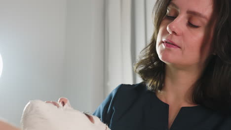 beauty technician conversing with female client while gently spreading thick face scrub from neck to forehead using soft silicone brush under bright lamp for calming spa treatment session