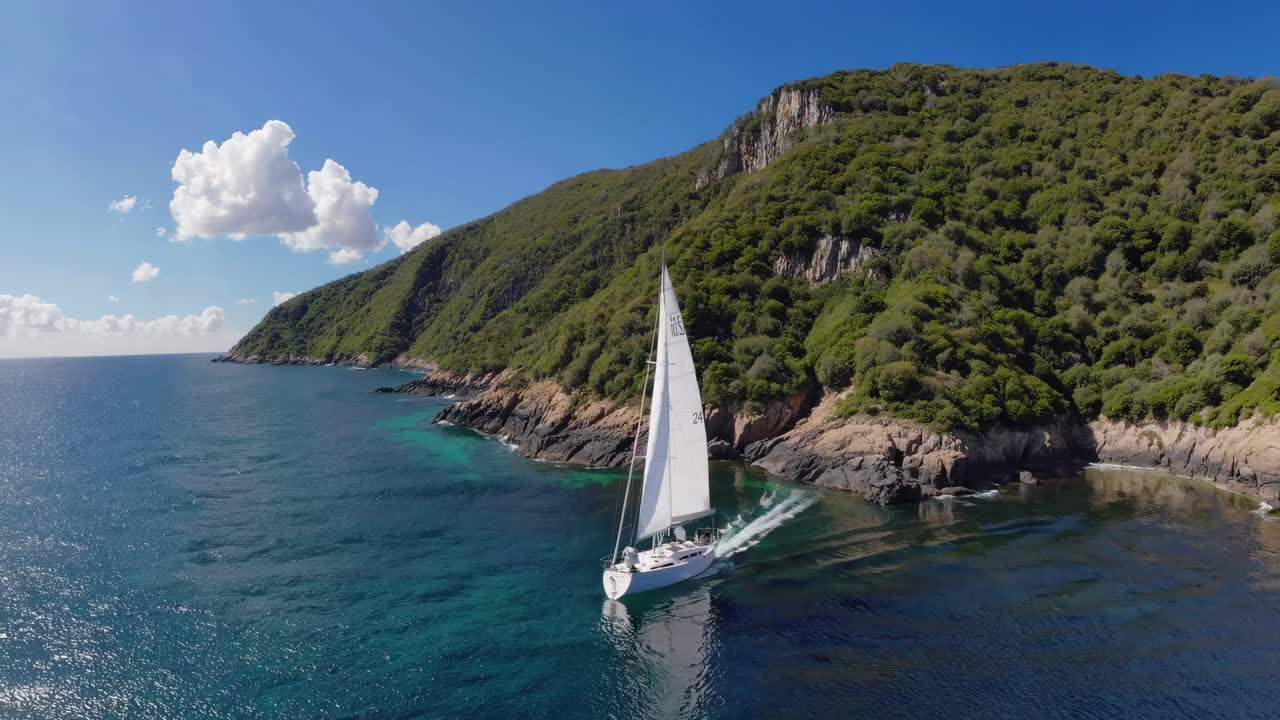 Aerial video captures a sailboat gliding along a lush, rocky coastline under a clear blue sky