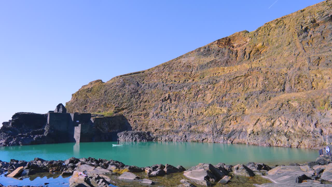 Deep Historic Blue Lagoon Quarry with Steep Sheer Cliff Edges with Old Buildings and Calm Sea Water with Surrounding Rocks.