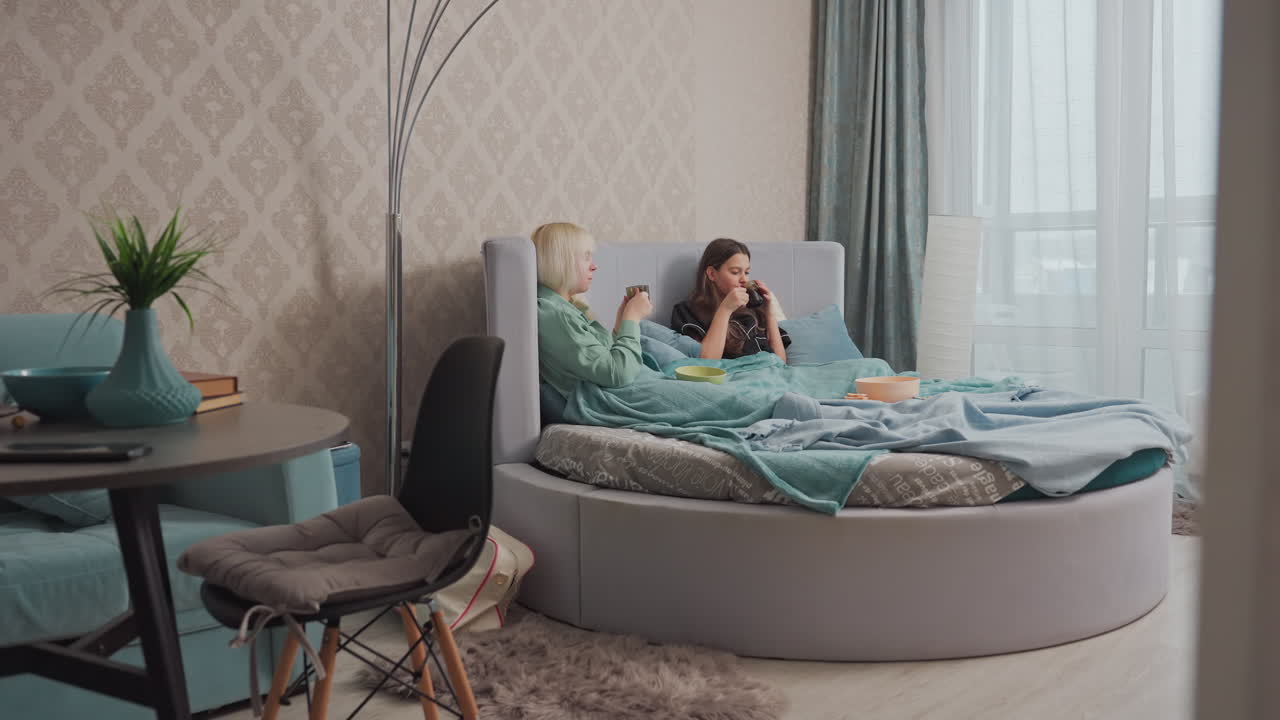 Two young female friends wrapped in blankets relax on bed while sipping coffee and chatting, soft morning light filters through window curtains as they enjoy warm drinks in peaceful bedroom