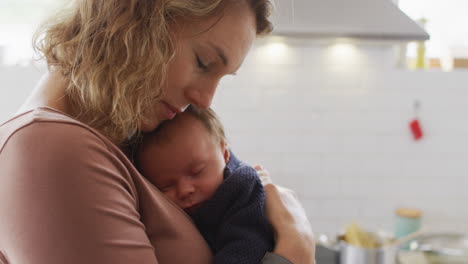 video of caucasian mother with newborn baby in baby carrier hugging in kitchen