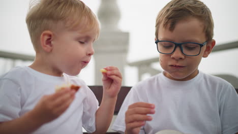 two siblings are seated on a bench, sharing chips, the younger child holds a snack in his right hand while the older sibling gives his younger brother some chips to enjoy