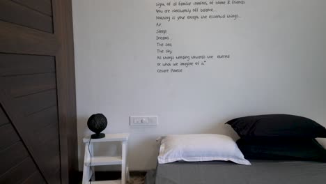 Minimalist-Bedroom-Interiors-With-Traveling-Quote-On-The-White-Wall-Above-The-Gray-Bed-With-Black-And-White-Pillows,-Small-Black-Lamp-On-The-White-Side-Table---tilt-down-shot