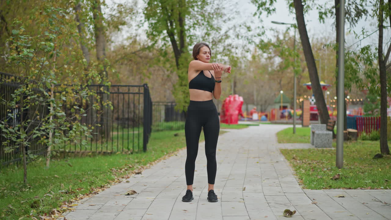 Young lady stretching arms outdoors in front of playground with game machines, iron rail fence, and round trees, creating a peaceful outdoor exercise setting in a game center environment