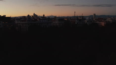 Ascending-footage-of-trees-in-park-silhouettes-against-colour-sunset-sky.-Revealing-evening-cityscape-at-dusk.