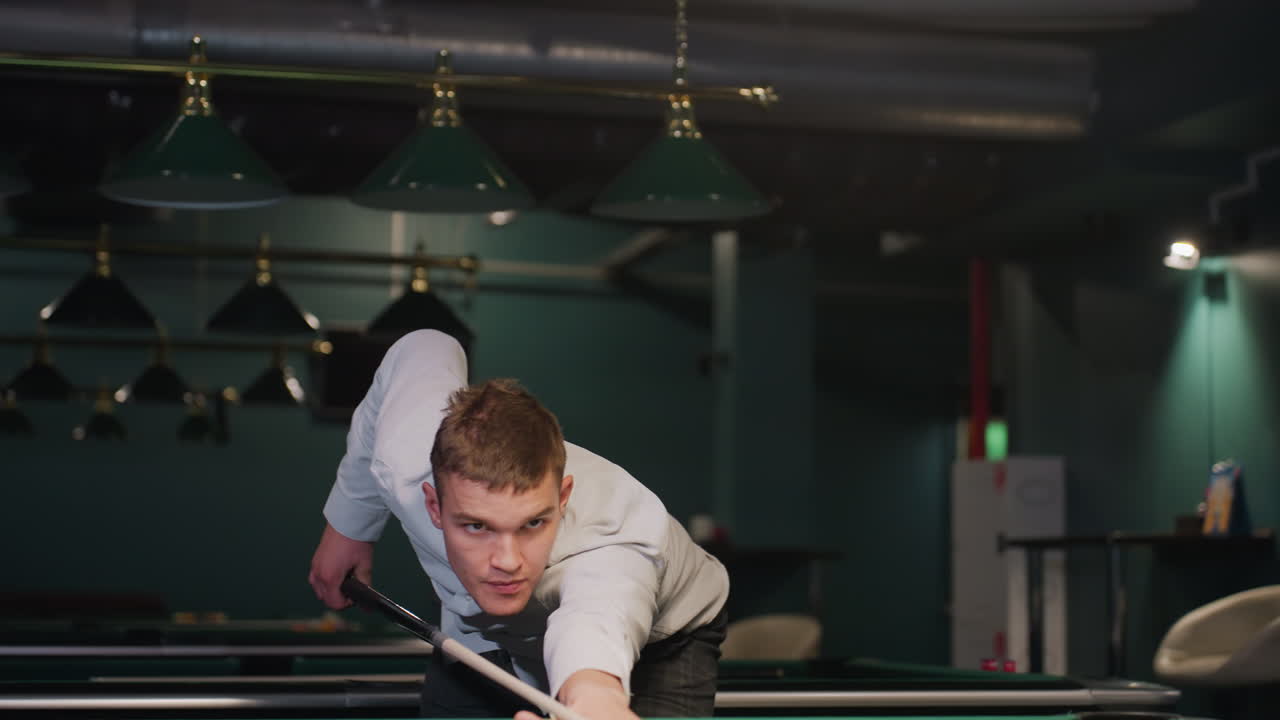 색 셔츠와 회색 바지를 입은 신사는 강렬한 초점을 맞추는 을 잡는 데 기대고 있습니다. 녹색 테이블과 오버헤드 조명이있는 희미하게 조명 된 빌리어드 홀은 극적인 분위기를 만니다.