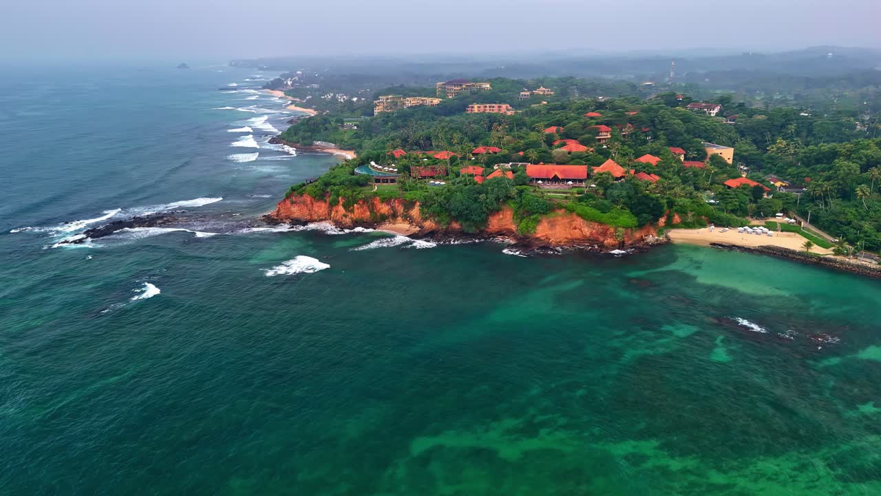 Sri Lanka'daki Weligama balıkçı köyü sahil şeridinin dron görüntüleri; kırmızı topraklı, palmiye ağaçları ve köy binalarıyla yemyeşil bir yarımadanın turkuaz denize uzandığını ve dalgaların kayalık kıyıya çarptığını gösteriyor.