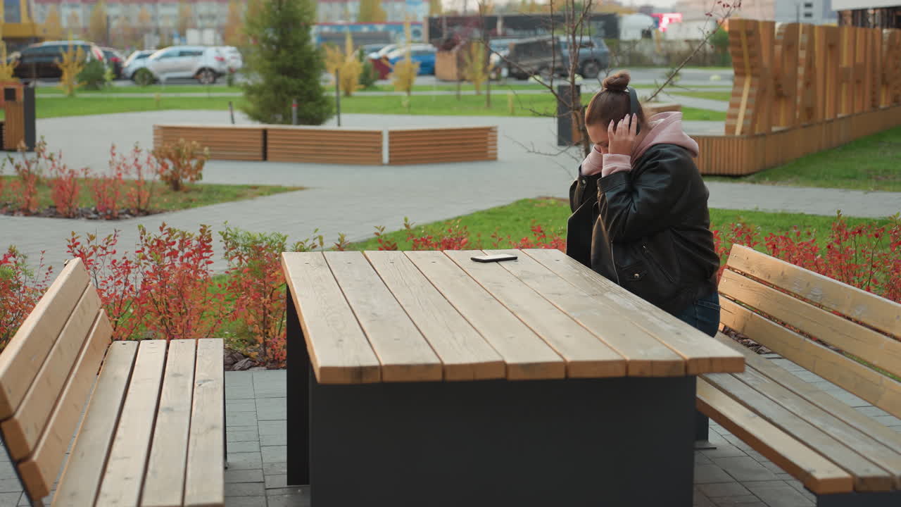 Student seated at wooden table in leather jacket wear headphones in outdoor park setting surrounded by red shrubs swaying gently in wind with benches, paved ground, and parked cars visible