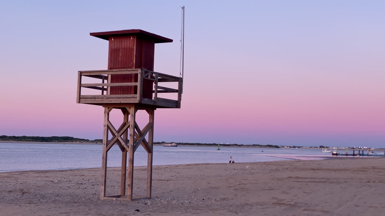 Torre de salvavidas vigila una tranquila playa de arena y aguas en calma durante un colorido atardecer en Sanlúcar de Barrameda