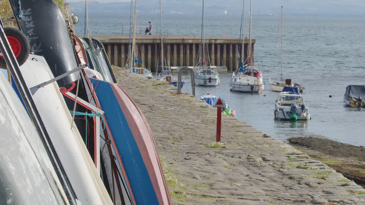 Camera slowly pans along stone dock with moored fishing boats in Scottish harbour, daylight