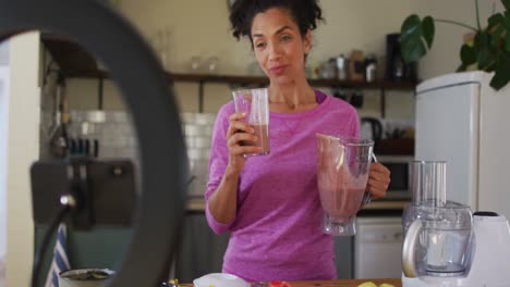 mixed race female vlogger drinking fruit juice and recording on smartphone in the kitchen at home