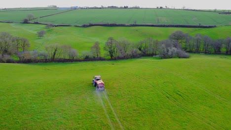 smooth aerial drone view following a red tractor fertilising a greet field with trees