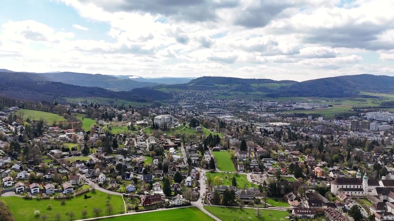 An aerial drone moves toward the Goetheanum in Dornach-Arlesheim, Switzerland, over homes, tree-lined streets and green fields, with wooded hills on the horizon beneath broken clouds.