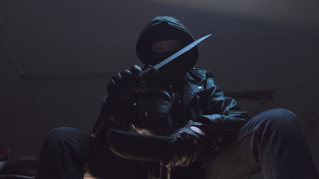 criminal con máscara de bandido y guantes de cuero negro inspecciona cuchillo