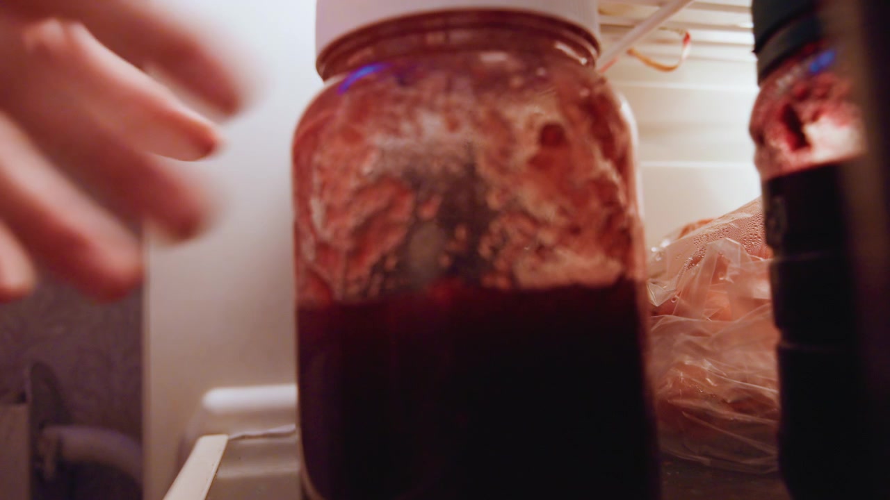 Mano busca tarro de mermelada en la nevera, cocinero casero inspecciona salsa en conserva en estante, movimiento de primer plano muestra cristal manchado, sobras envueltas en plástico cerca, luz cinematográfica granulada y gesto táctil.