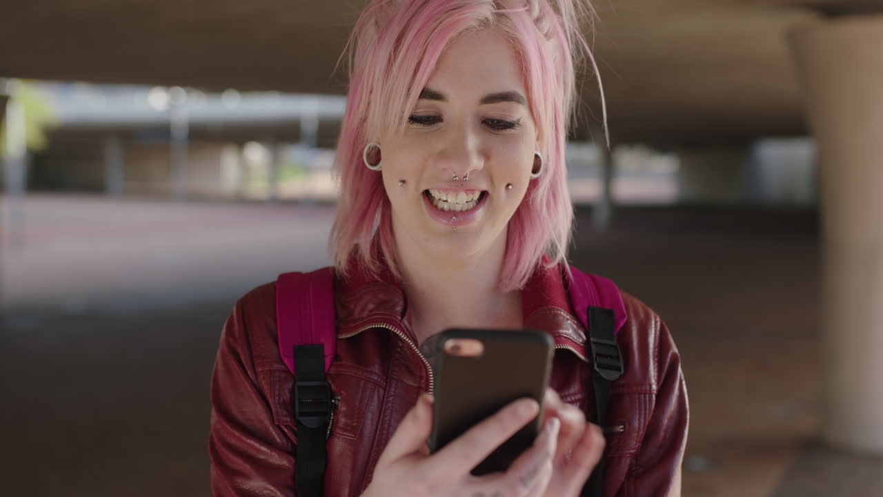 retrato de una mujer punk alternativa con cabello rosa enviando mensajes de texto navegando por las redes sociales usando un teléfono inteligente fondo urbano