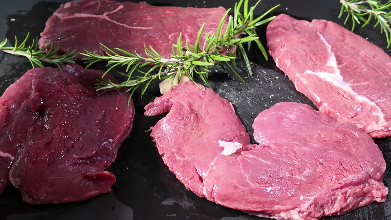 Close-up of raw beef steaks being seasoned with salt by hand, featuring fresh rosemary and garlic on a dark cooking surface