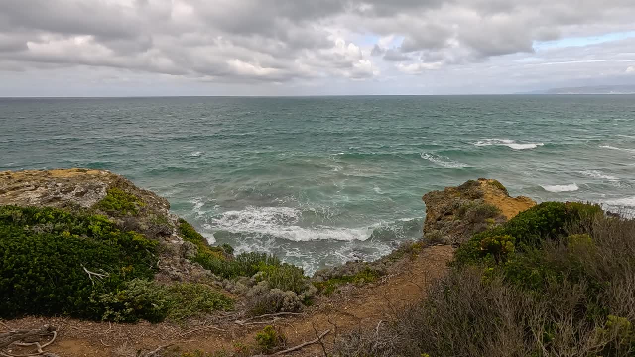 Waves surge against rugged cliffs and coastal vegetation under cloudy skies. Static wide shot captures dynamic ocean movement and dramatic natural landscape along Great Ocean Road