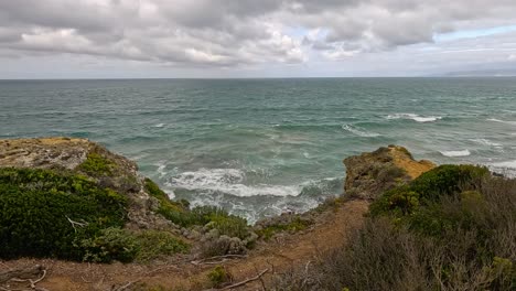 waves surge against rugged cliffs and coastal vegetation under cloudy skies. static wide shot captures dynamic ocean movement and dramatic natural landscape along great ocean road