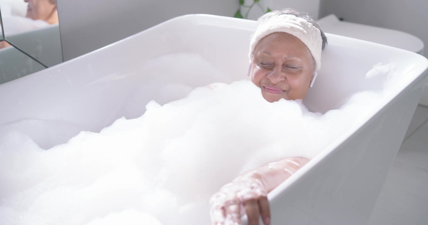 Un'anziana donna afroamericana ascolta musica per rilassarsi tramite auricolari, raccogliendo la schiuma nella vasca da bagno.
