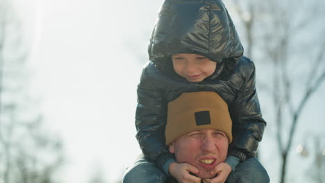 primer plano de un padre llevando a su hijo sonriente en sus hombros, ambos disfrutando de un momento de alegría, las manos del niño descansan bajo la cara del padre, con suaves rayos de luz reflejados por detrás