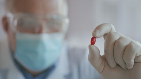 Extreme-close-up-of-scientist-in-the-mask-developer-of-the-vaccine-against-coronavirus-holds-in-his-hands-the-red-pill.-The-doctor-looks-at-the-painkillers-antiviral-medication.-Vitamins.-High-quality-4k-footage