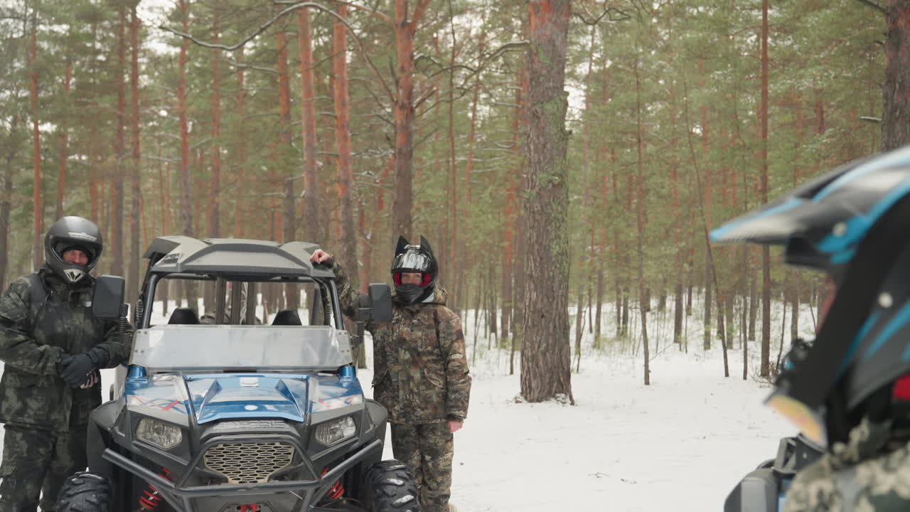 Corsa estrema in quad nella foresta innevata, due piloti con casco accanto a un UTV blu in un bosco di pini, giacche mimetiche e occhiali, terreno innevato, primo piano del volto del pilota attraverso la visiera, mani sull'acceleratore, in preparazione.