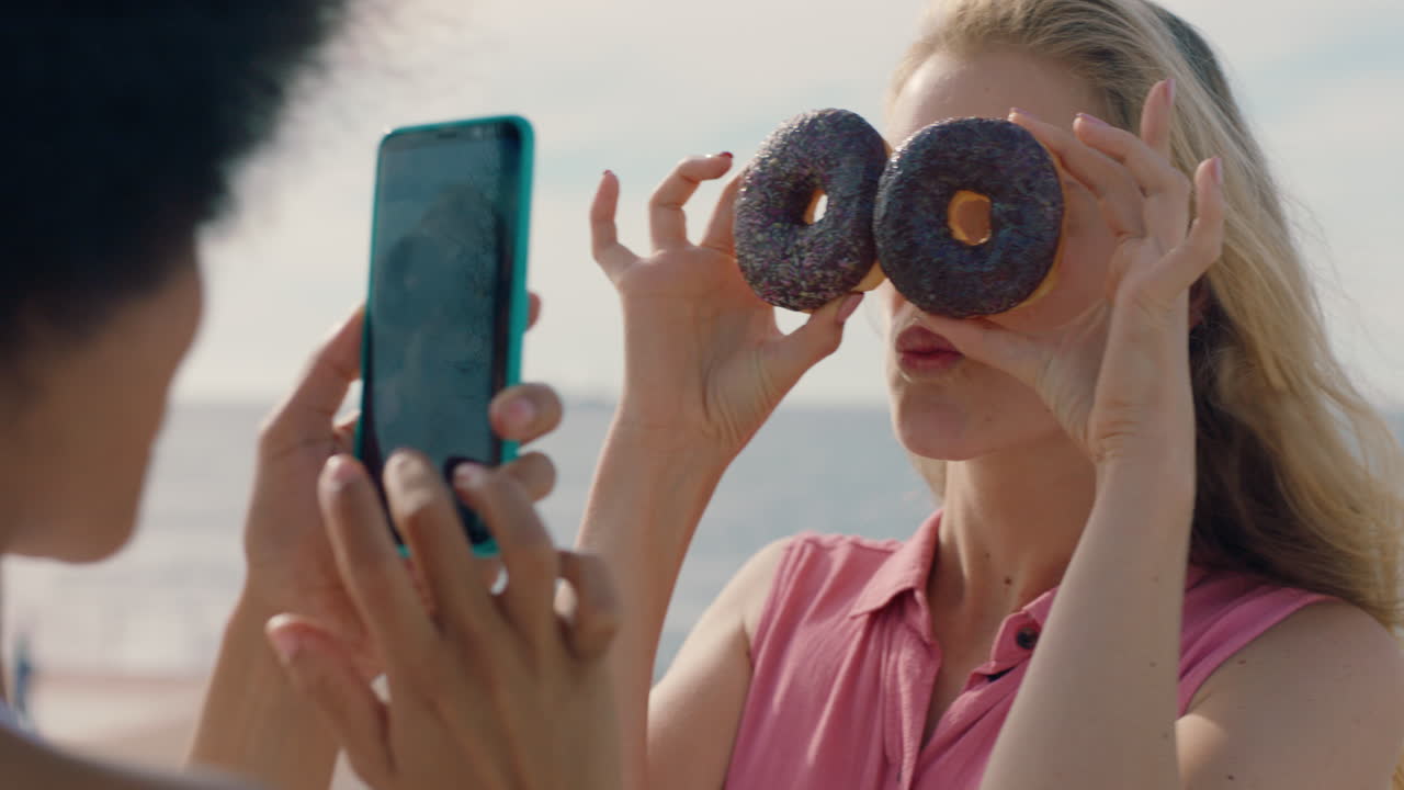 hermosa mujer posando con rosquillas en la playa mejor amiga tomando fotos usando teléfono inteligente compartiendo fin de semana en la playa en las redes sociales disfrutando de la diversión de verano 4k
