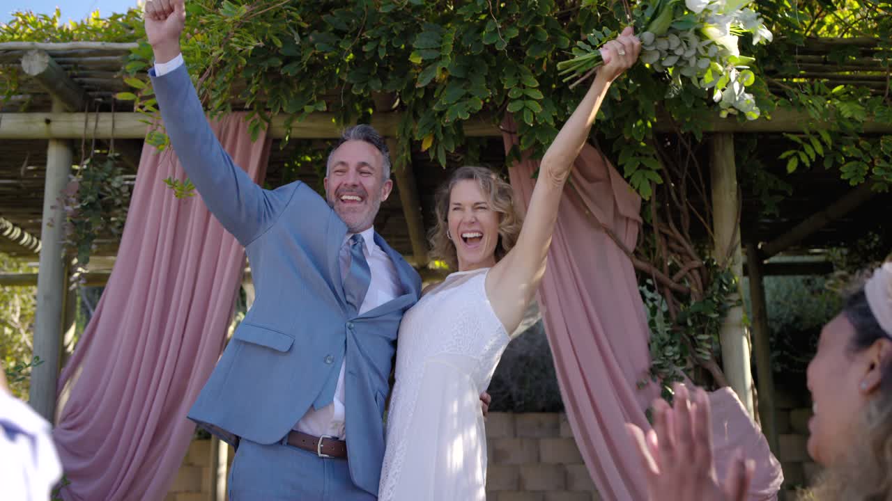 Coppia di sposi novelli in abito da sposa e completo che celebrano i voti nuziali vicino a un pergolato, alzando il bouquet e salutando gli invitati.