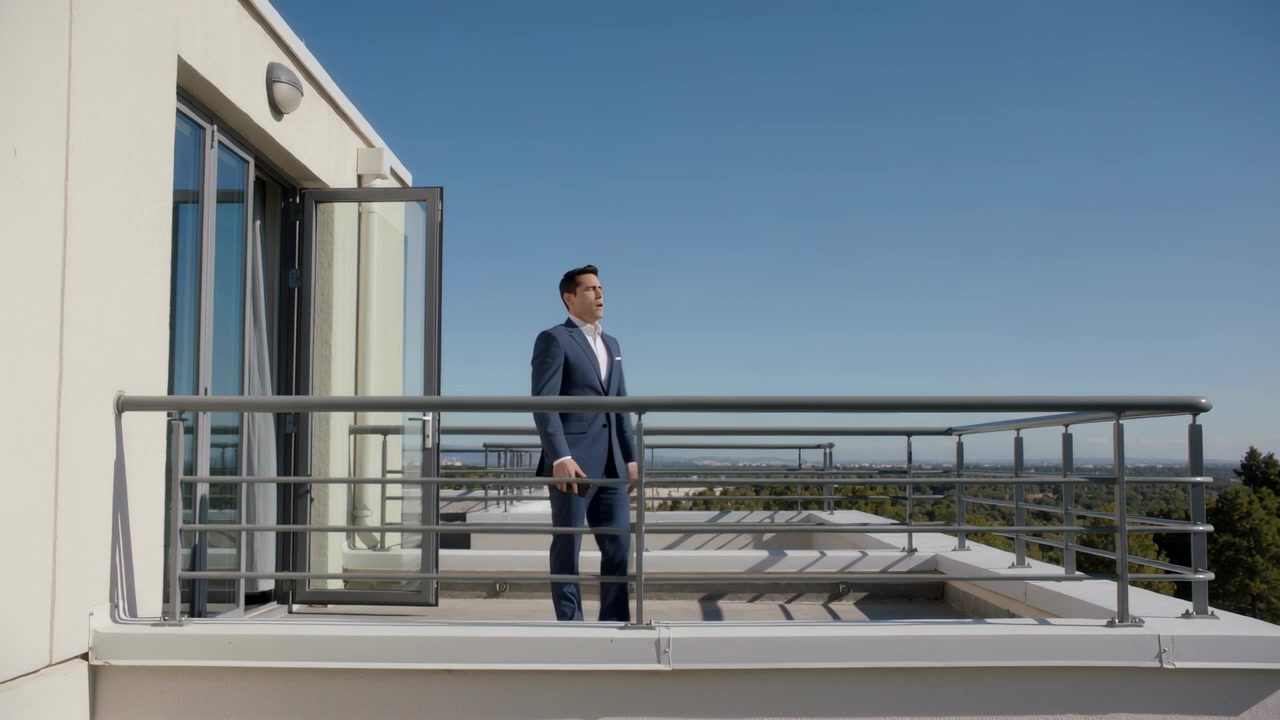 Puerta de cristal abriéndose, un hombre con traje azul marino saliendo al tejado, bajando el smartphone, observando el horizonte.
