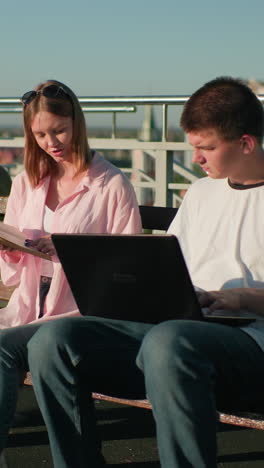 dos adultos jóvenes estudiando al aire libre, el macho en la computadora portátil mientras la hembra lee el libro y señala los detalles, en un paisaje urbano con edificios modernos y vegetación exuberante