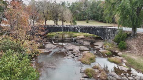 Romántico-Puente-De-Piedra-Sobre-El-Río-Con-Rocas-Y-Agua-Y-árboles-A-Los-Lados-Alejándose