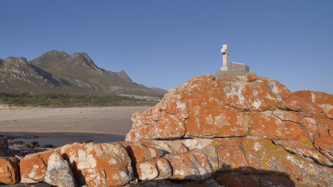 Ampia inquadratura di una croce su una costa rocciosa che si affaccia su acqua che scorre, la spiaggia e un piccolo insediamento umano ai piedi di una montagna in lontananza, a Kleinmond, Sudafrica.