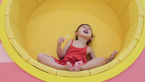 Caucasian-Blonde-Girl-Eating-Delicious-Sweet-Yellow-Lollipop-Sitting-in-Circular-Wall-Built-in-Chair---low-angle-view