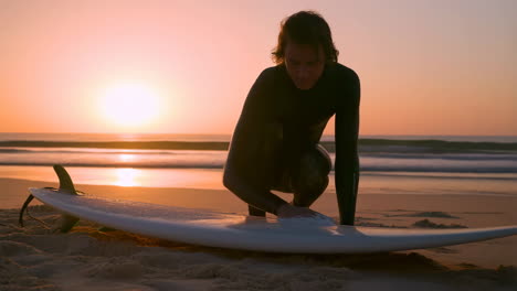 surfer waxing board on beach at sunset