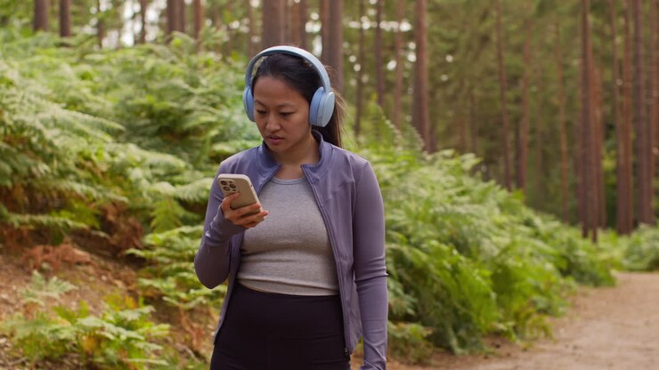 Donna che indossa cuffie wireless, si allena all'aperto ascoltando musica o podcast in streaming dal telefono cellulare, correndo lungo un sentiero nella foresta e indossando abbigliamento sportivo