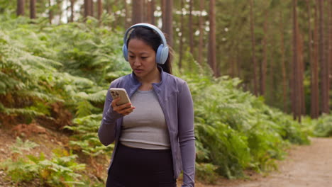 woman wearing wireless headphones exercising doing work out outdoors streaming music or podcast from mobile phone running along track in forest wearing sports clothing