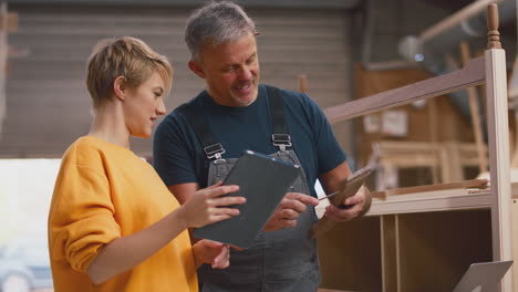 Female-Apprentice-Learning-From-Mature-Male-Carpenter-With-Digital-Tablet-In-Furniture-Workshop