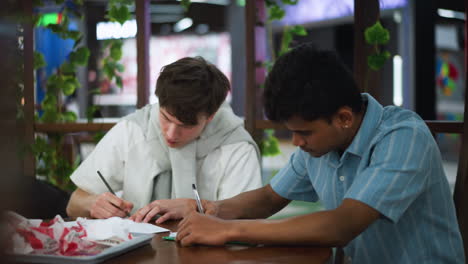 camera capturing two determined friends making sketches on paper while wooden pillar blocks part of frame close up focus on hands drawing creative collaboration in workspace with warm light softly l