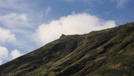 a-lone-hiker-stands-and-surveys-the-view-from-the-top-of-an-Oahu-outcrop-with-blue-skies-and-clouds-behind