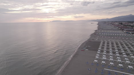 Un-Dron-Disparó-Sobre-Las-Playas-De-Viareggio,-Italia,-Durante-La-Puesta-De-Sol-En-Un-Día-Gris-Volando-Hacia-El-Mar-Con-Edificios-Y-Montañas-En-El-Fondo.