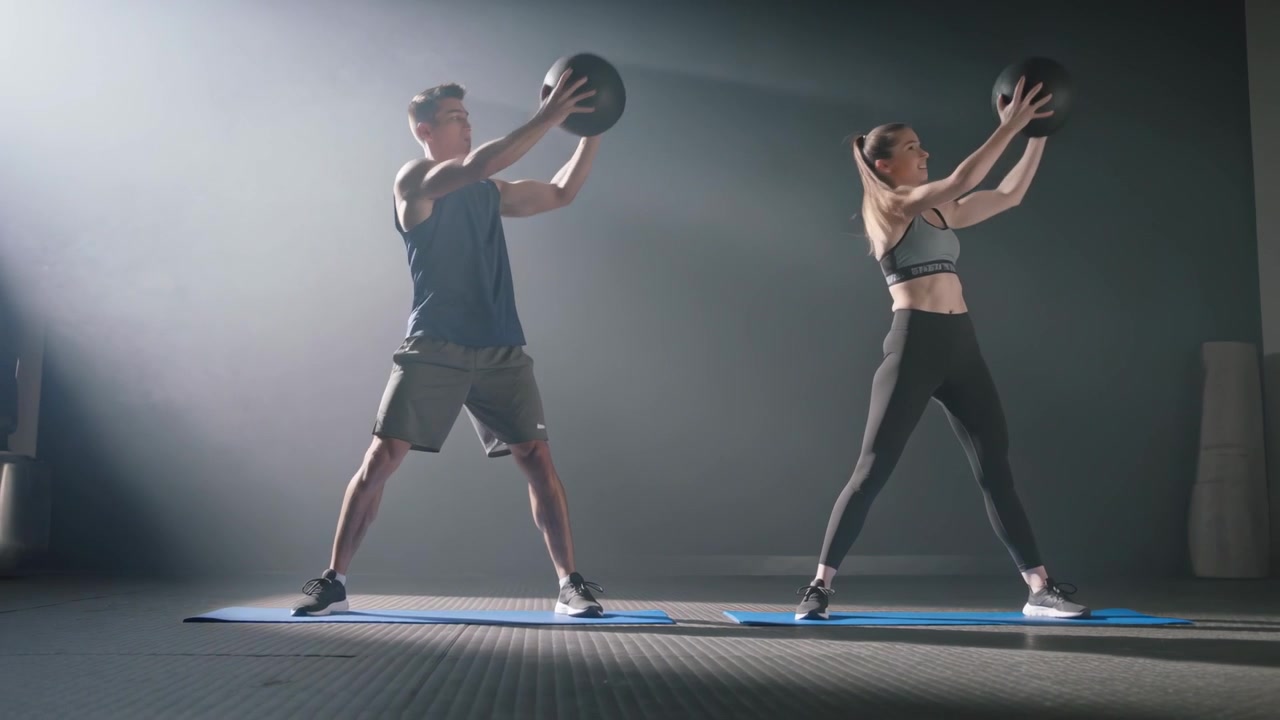 Reacting to cue, trainers in-gymwear doing medball lunges on blue mats in-gym-studio for training