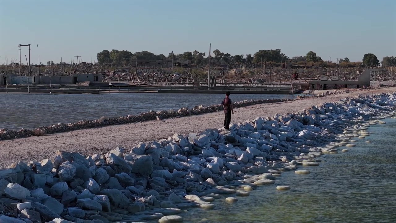 Villa Epecuen'in tarihi sel kalıntıları arasında, ıssız bir yolun kenarındaki kayalık kıyıda duran yalnız turist. Güneşli ve rüzgarlı bir öğleden sonra alçak havadan yörünge çekimi.