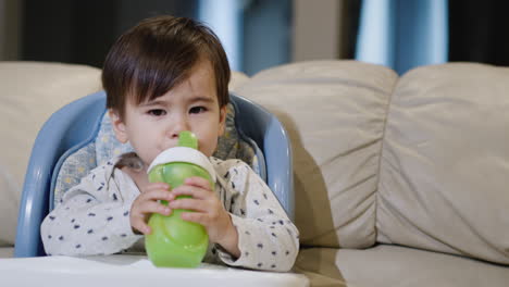 Ein-Zweijähriges-Baby-Trinkt-Milch-Aus-Seiner-Flasche,-Sitzt-Auf-Einem-Hochstuhl